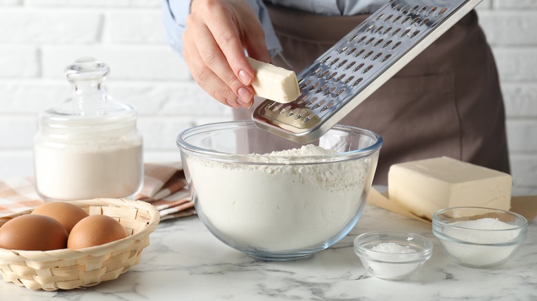 Hands grating butter with a cheese grater into bowl of flour