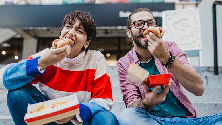 Two people eat hot dogs while sitting on the stairs